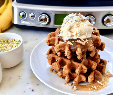 Stack of waffles topped with ice cream in the foreground, with bowl of sprinkles, bananas, and GreenPan waffle maker in the background